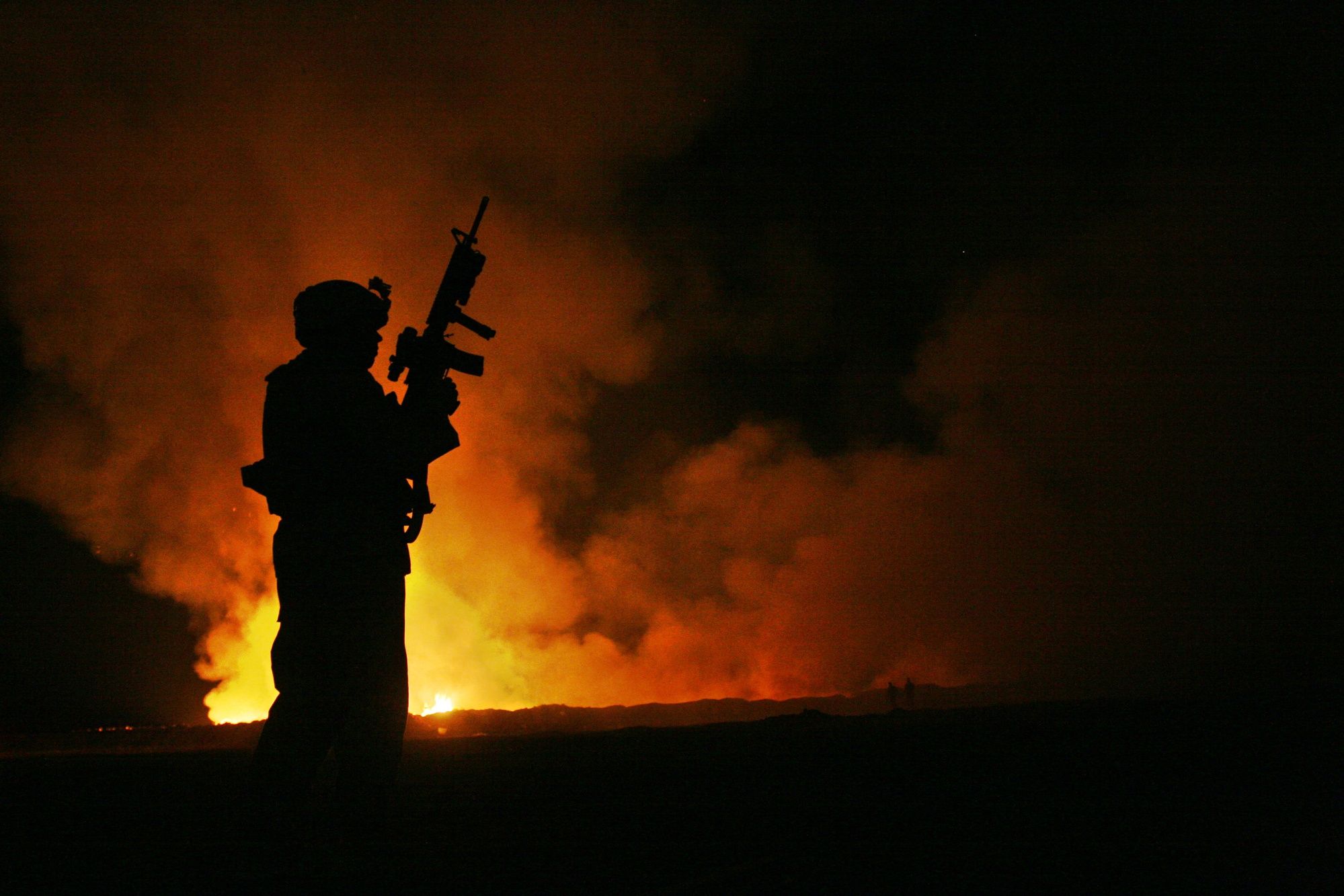 The dark silhouette of a service member stands in front of smoke and flames rising from a burn pit at night.