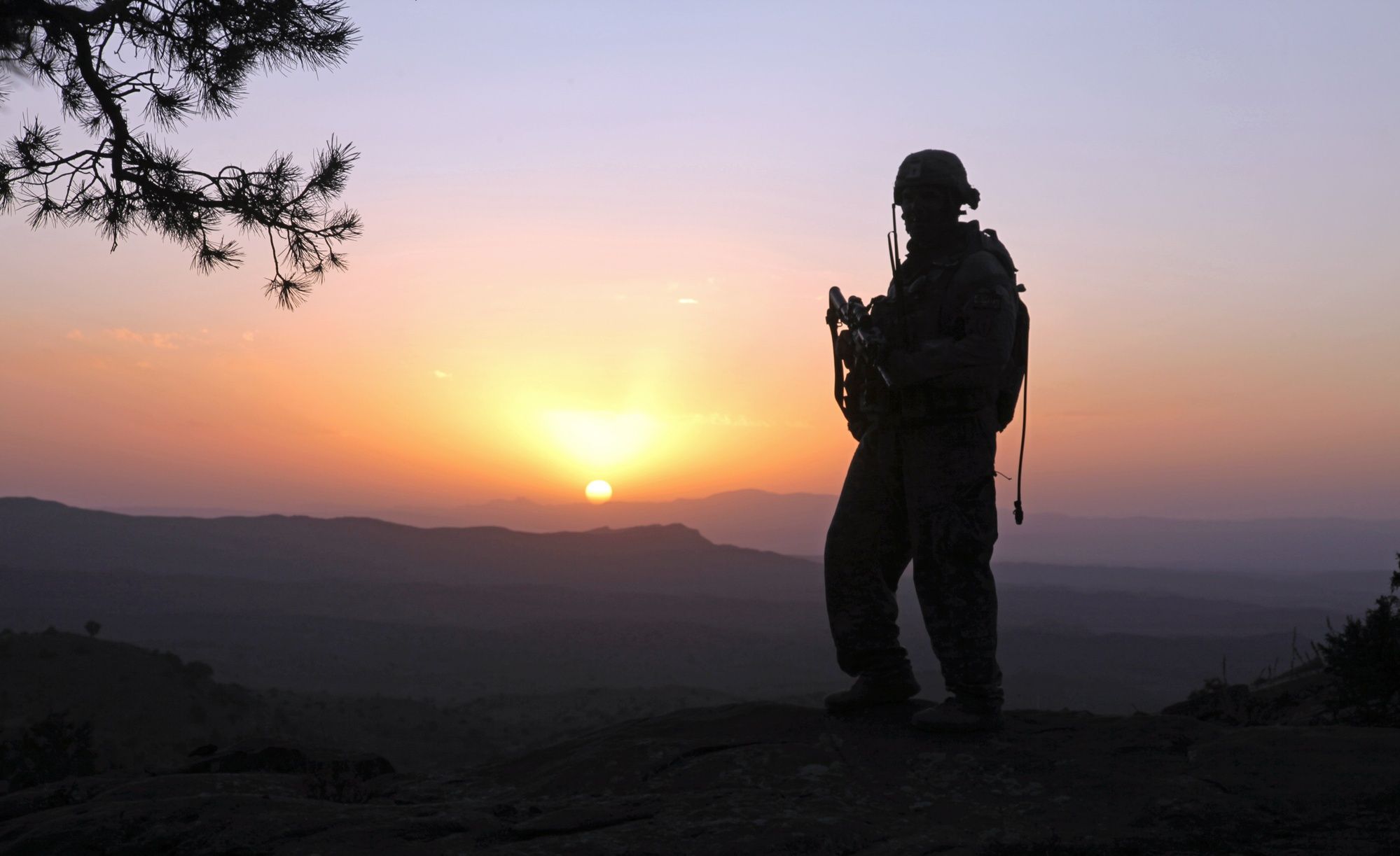 The silhouette of a soldier in full gear stands on a ridge as the sun sets over distant mountains.