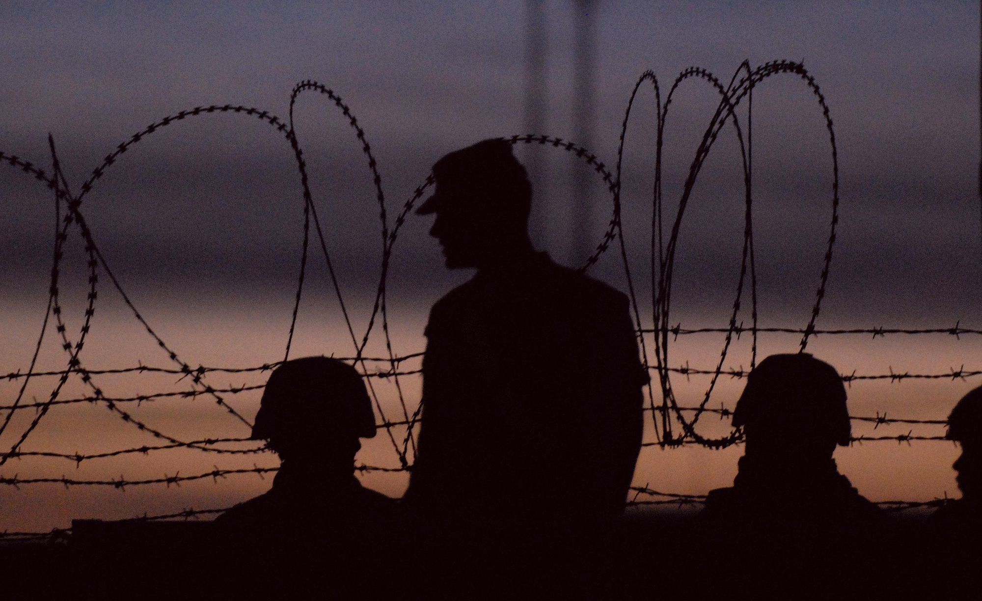 Silhouettes of helmeted soldiers behind coils of concertina wire against a dusk sky.