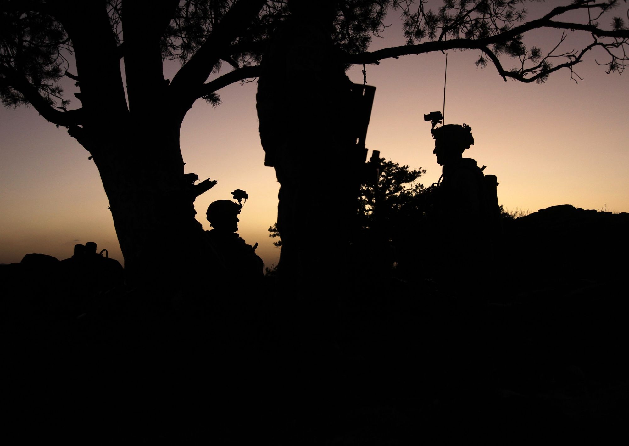Silhouettes of soldiers resting beneath a tree against an orange sunset sky.