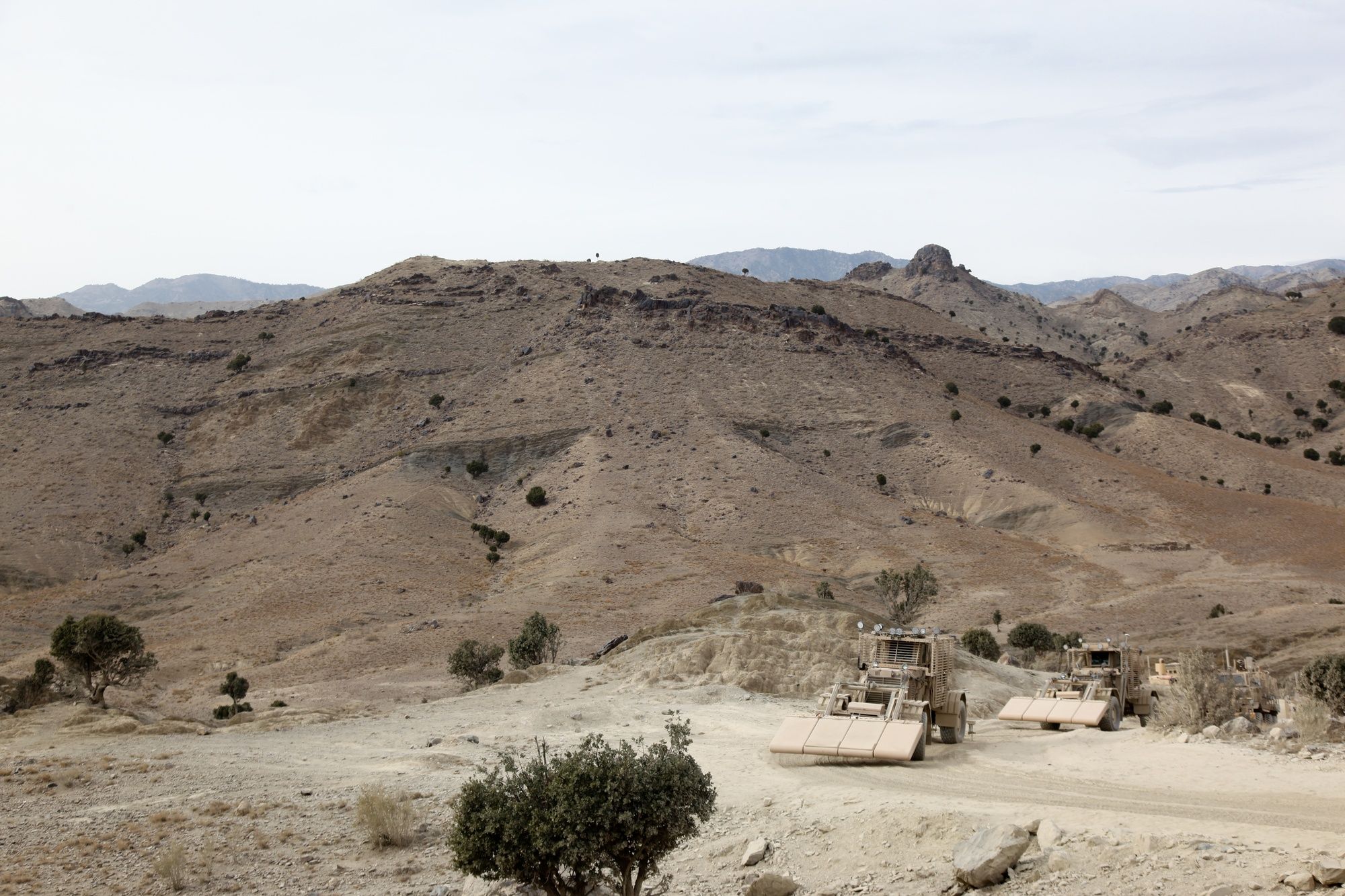 Route-clearance vehicles travel a dirt road through a wide, dry mountain valley.