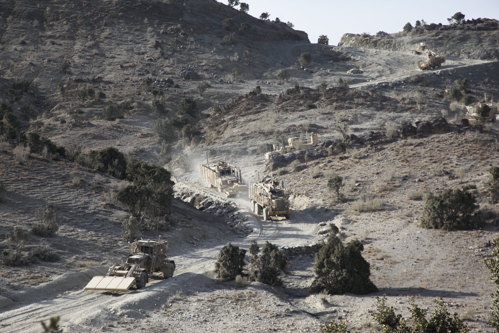 A line of armored route-clearance vehicles kicks up dust as it climbs a winding mountain road.