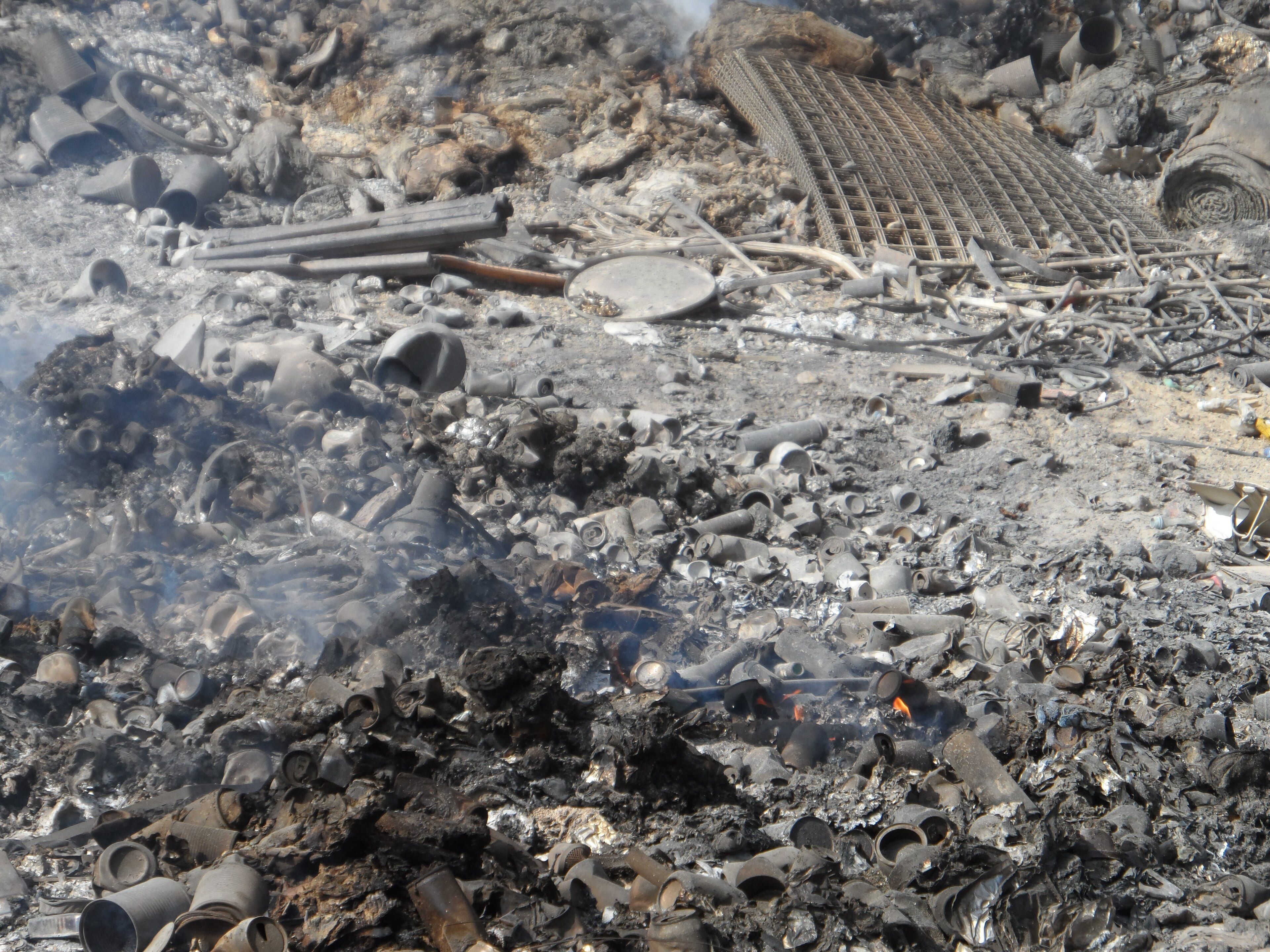 Smoke rises from a field of charred trash and debris in an open-air burn pit.