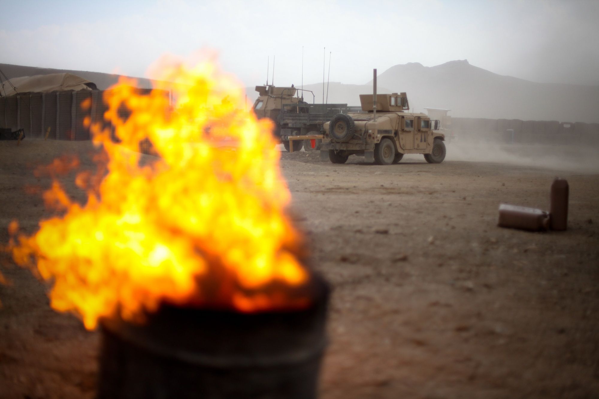 Flames leap from a burn pit in the foreground while armored vehicles sit in the dust behind it.