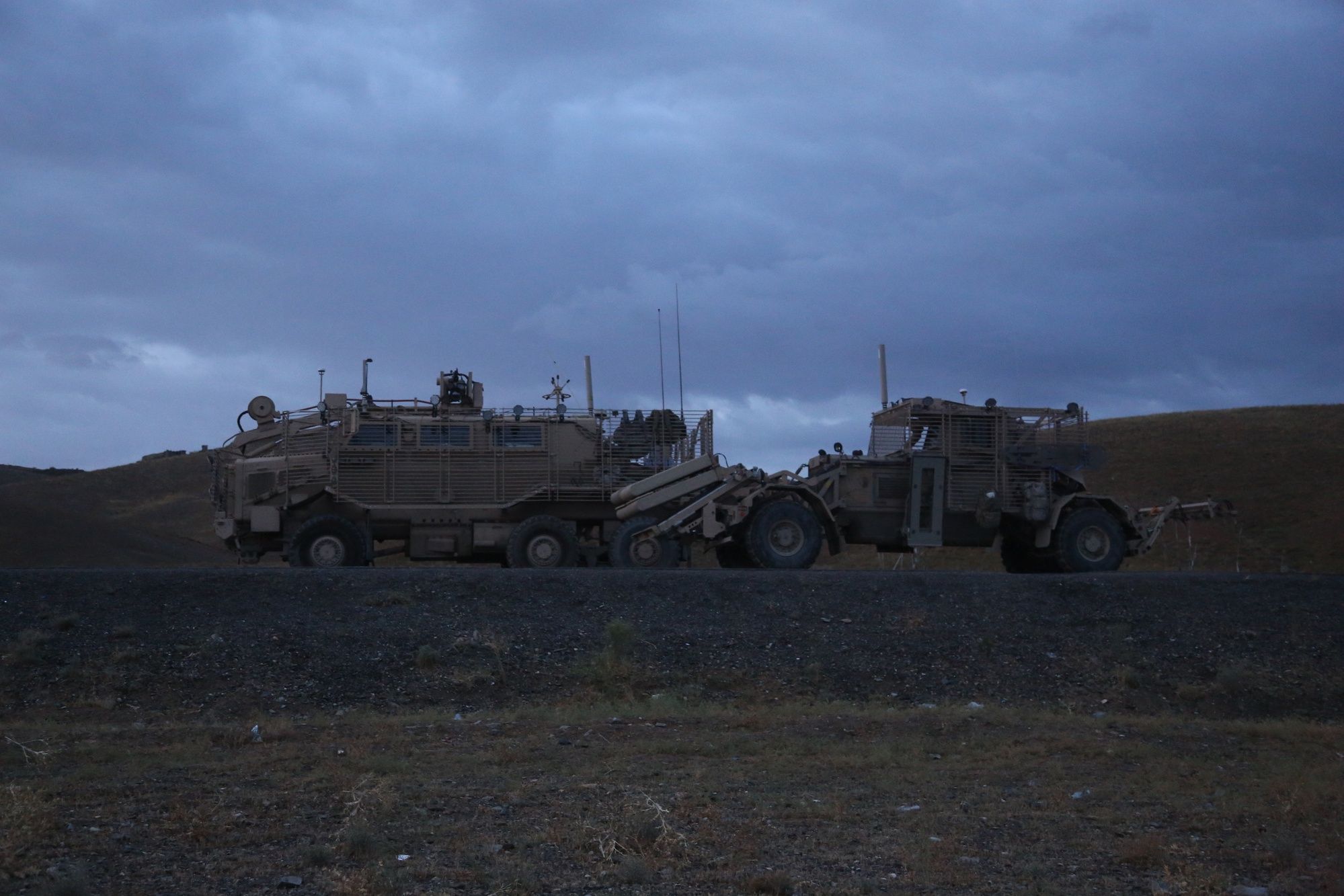 Armored route-clearance vehicles parked on open ground under a dark, stormy dusk sky.