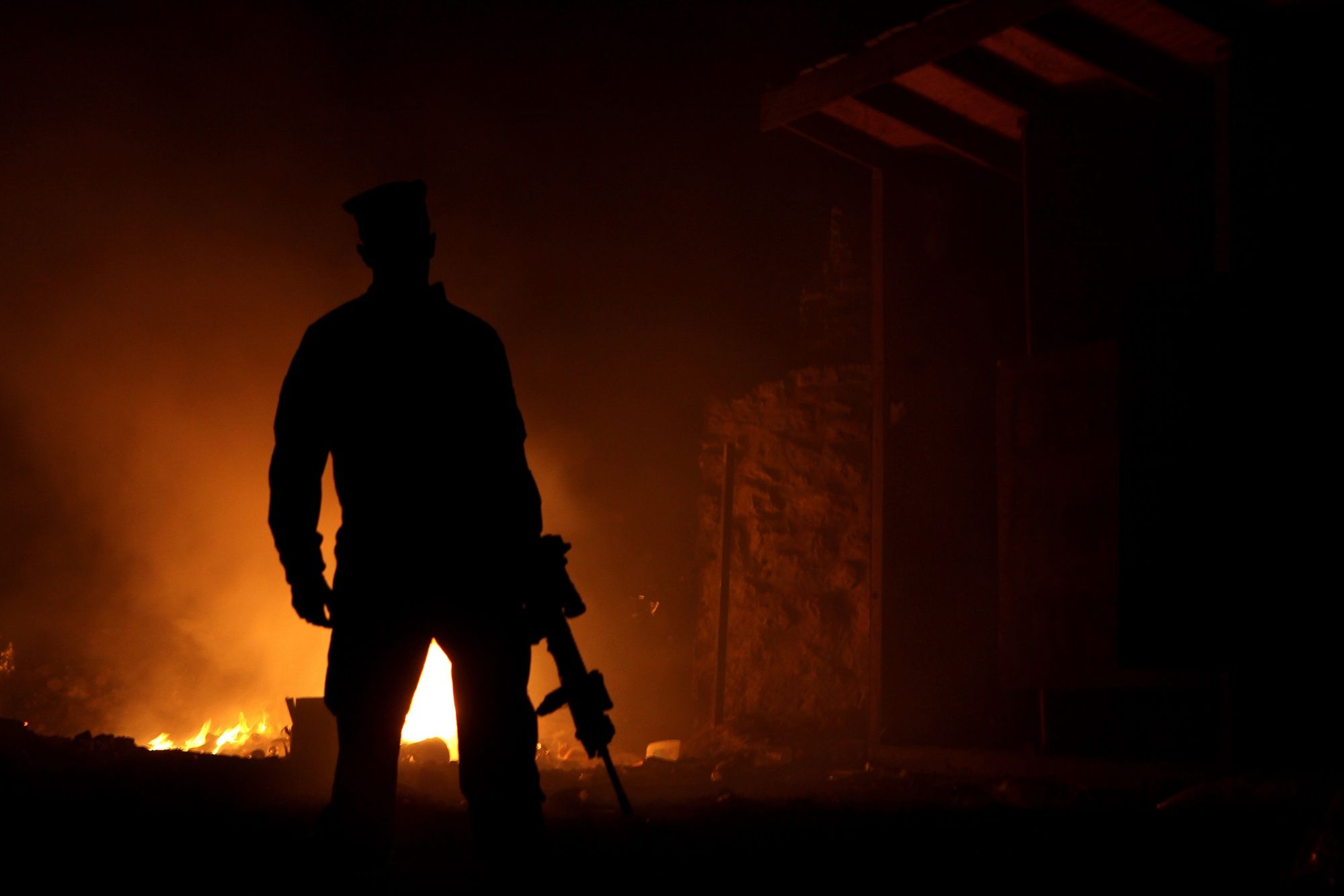 The dark silhouette of a Marine stands before the orange glow of a burn pit at night.